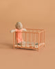 A small stuffed doll in a pink outfit stands inside the Maileg Playpen, Micro - Dark powder, featuring a patterned mattress and set against a plain peach background.