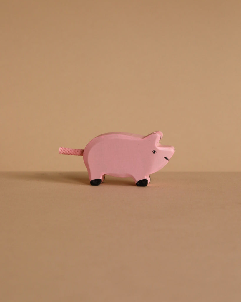 A pink Holztiger Handmade Wooden Farm Animals Set pig with a rope tail and black-painted feet.