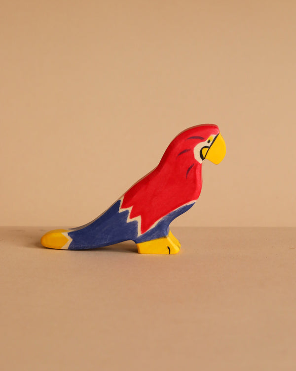 The Holztiger Parrot, a small handcrafted wooden figurine painted red, blue, yellow, and white, is displayed on a neutral beige surface against a plain beige background. (Ships in one week.).
