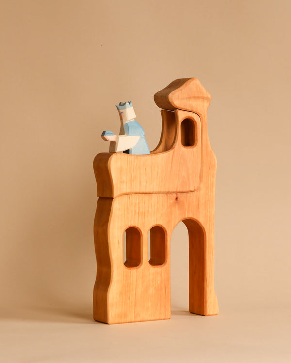 A handcrafted wooden toy figure sits atop the Ostheimer Lookout - Large, a castle-shaped wooden block structure. The toy figure, resembling a king or royal, is adorned with a blue crown and a blue and white outfit. The Ostheimer Lookout - Large features an arched doorway and two window-like openings, ideal for sparking imaginative play.