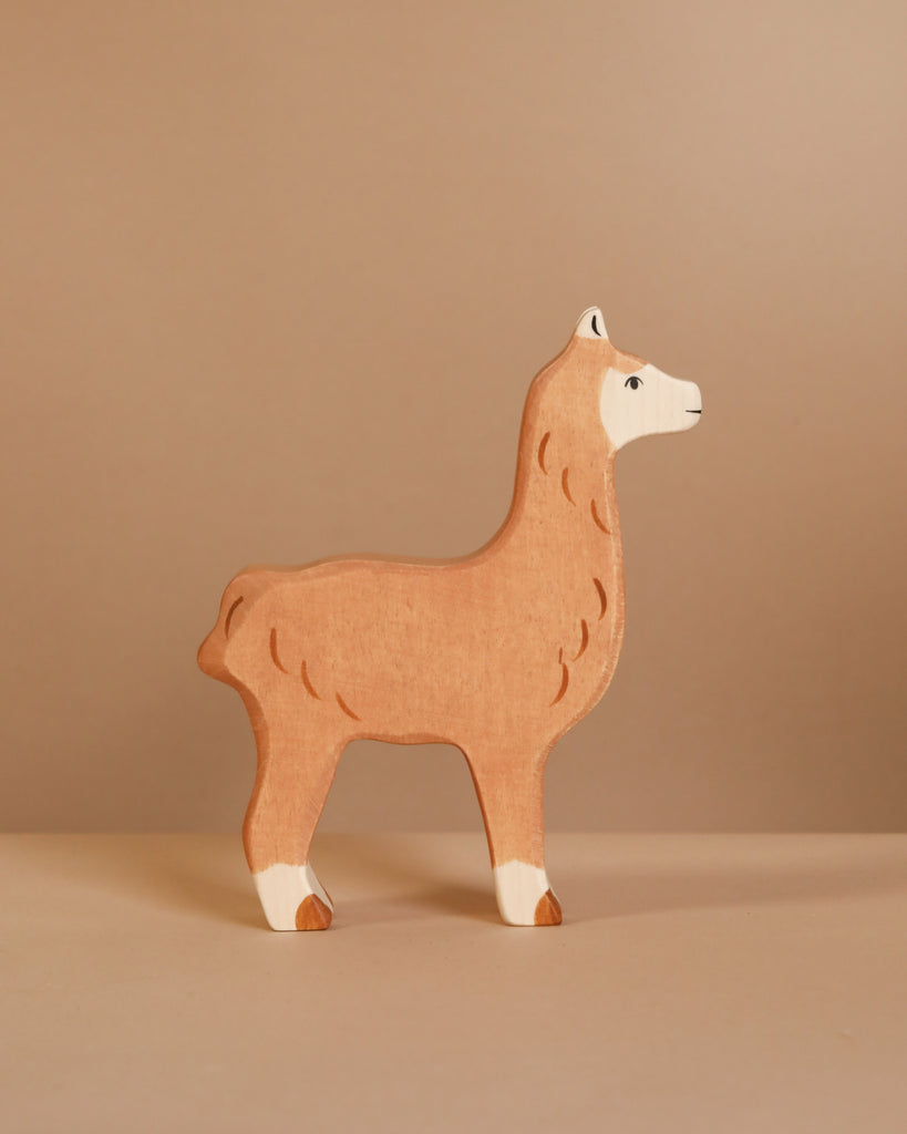 A light brown, white-accented llama from the Holztiger Handmade Wooden Farm Animals Set stands on a beige surface with a matching background.