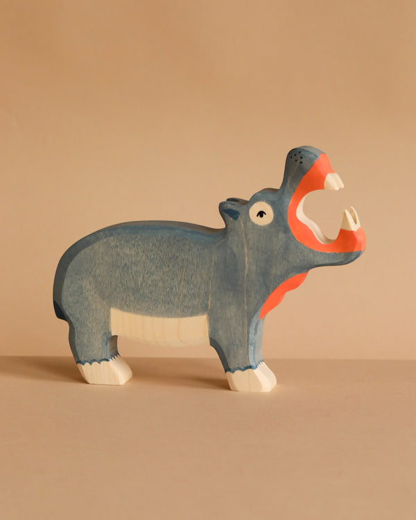 The Holztiger Hippo, a handcrafted wooden figurine painted blue with an open mouth showing white teeth and pink insides, stands on a beige surface against a matching background. Ships in one week.