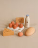 A display of Sabo Concept Handmade Wooden Dairy Set items that includes eggs, cheese, milk, and butter on a beige background, designed to look realistic with non-toxic paint.