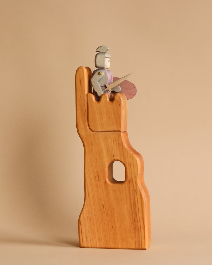 An Ostheimer Lookout - Small wooden toy knight stands atop a stylized alder wood castle turret. The knight holds a sword and shield and wears a simple helmet. The turret has a rectangular window cutout in the middle. The background is a neutral light brown, highlighting the beauty of handcrafted wooden toys.