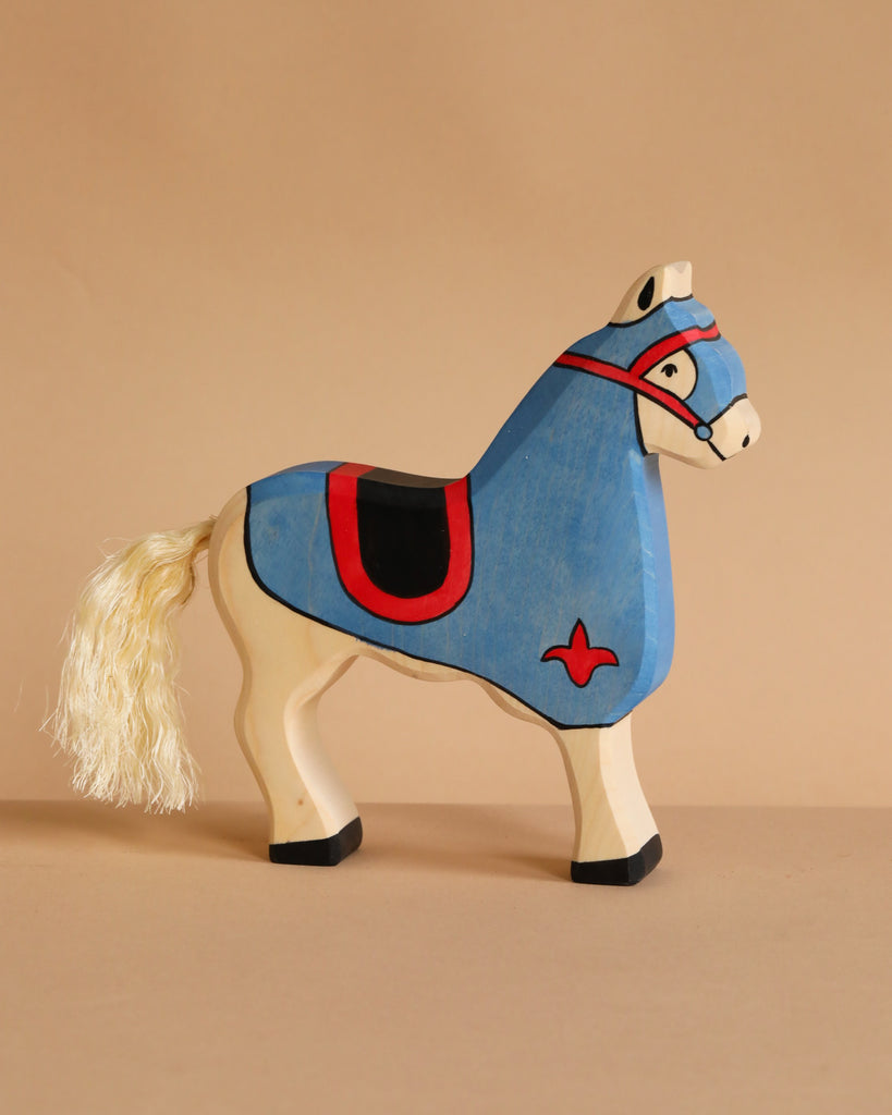 The Holztiger Tournament Horse, Blue is a handcrafted wood toy featuring a cream body, blue saddle and headpiece, red and black accents, and a stringy tail. Inspired by HOLZTIGER figures, it rests on a plain light brown surface.