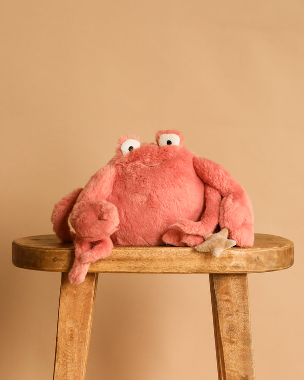 The Mon Ami Caldwell Crab Plush Toy, featuring big googly eyes and a soft pink exterior, rests atop a wooden stool against a beige background—a charming keychain plush or bag accessory.