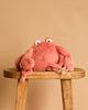The Mon Ami Caldwell Crab Plush Toy, featuring big googly eyes and a soft pink exterior, rests atop a wooden stool against a beige background—a charming keychain plush or bag accessory.