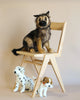 A realistic German Shepherd Puppy Stuffed Animal sits on a wooden chair, looking forward, with a toy Dalmatian and a small toy dog with a red collar standing next to it on the floor.
