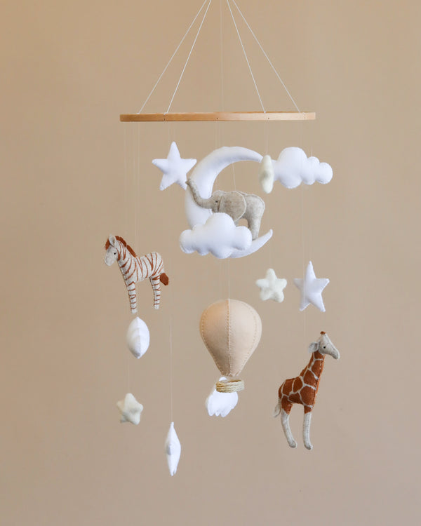 A Day Dreamer handmade mobile featuring plush animals including a zebra, elephant, and giraffe, alongside clouds, stars, and a hot air balloon, all hanging against a soft beige background.