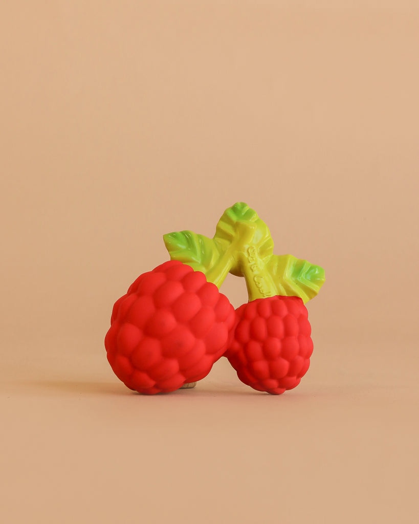 Valery the Raspberry Teething Toy is a brightly colored sensory teether made of natural rubber, shaped like two raspberries with green leaves, displayed on a plain, peach-colored background.