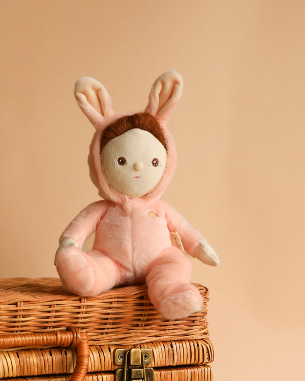 A soft, plush toy rabbit with human-like features, Olli Ella | Dinky Dinkums - Bella Bunny, sits on a wicker basket. The rabbit is a pastel peach color with floppy ears lined with