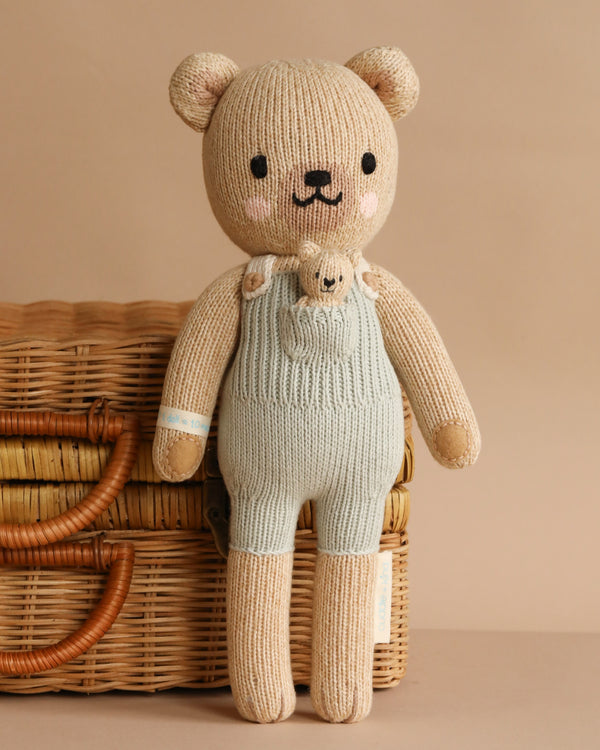Cuddle + Kind Alexander The Honey Bear, a handmade knitted teddy in light blue overalls, stands before a wicker basket. It has embroidered eyes, nose, and smile and is filled with hypoallergenic polyfill for softness.