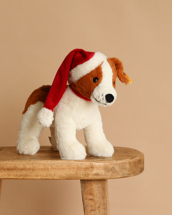 The Steiff Snuffy Puppy Dog Plush with Santa Hat for Holiday stands on a wooden stool against a plain beige background, making it an ideal festive gift.