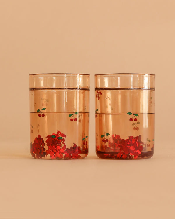 The Konges Sloejd Glitter Cups - Cherry (2 Pack) feature clear cups with red and green cherry designs, filled with water and red flower confetti—fun dinnerware to add a playful touch to any kids cup collection.