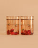 The Konges Sloejd Glitter Cups - Cherry (2 Pack) feature clear cups with red and green cherry designs, filled with water and red flower confetti—fun dinnerware to add a playful touch to any kids cup collection.