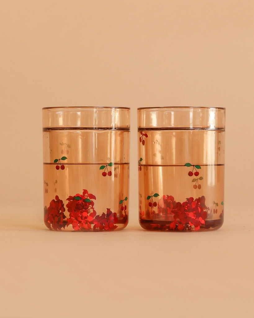 The Konges Sloejd Glitter Cups - Cherry (2 Pack) feature clear cups with red and green cherry designs, filled with water and red flower confetti—fun dinnerware to add a playful touch to any kids cup collection.