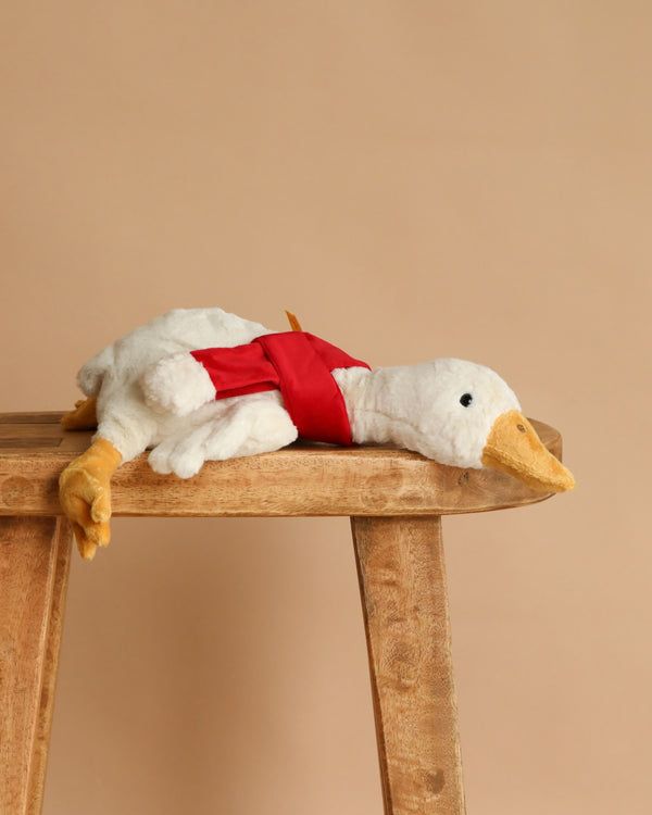 The Steiff Gilda Christmas Goose with Red Scarf rests softly on a wooden bench against a beige background—an ideal cuddle companion or delightful holiday gift.