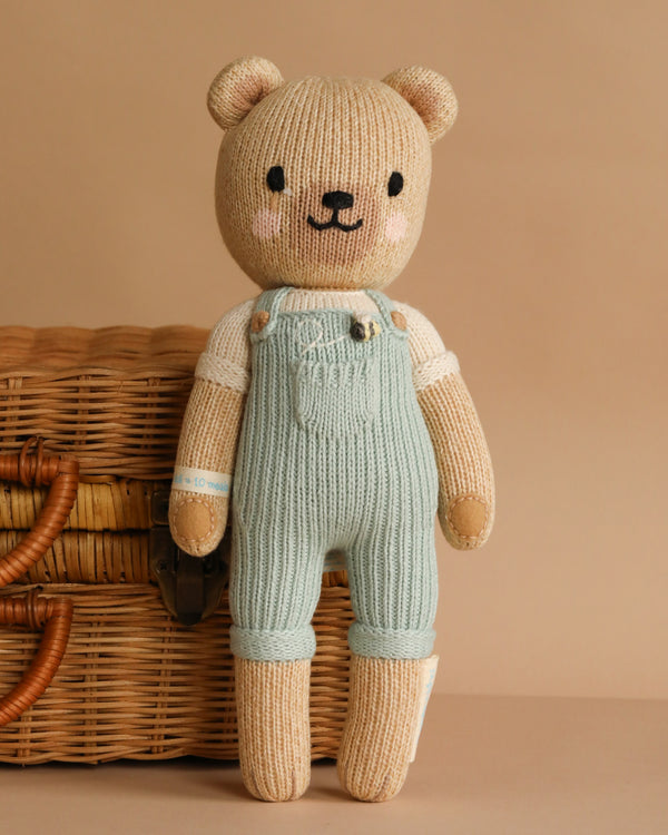 Cuddle + Kind Charlie The Honey Bear stands in light blue overalls and a cream shirt on a beige surface before a wicker basket, set against a plain beige backdrop. Hand-knit with eco-friendly materials and crafted using sustainable fair trade practices.