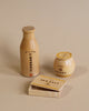 A photo of three wooden containers from the Milton & Goose Pantry Play Food Set placed on a beige background. On the left is a bottle labeled "Apple Cider Vinegar," in the middle is a jar labeled "Manuka Honey," and at the bottom is a box labeled "Sea Salt" with its lid slightly open.