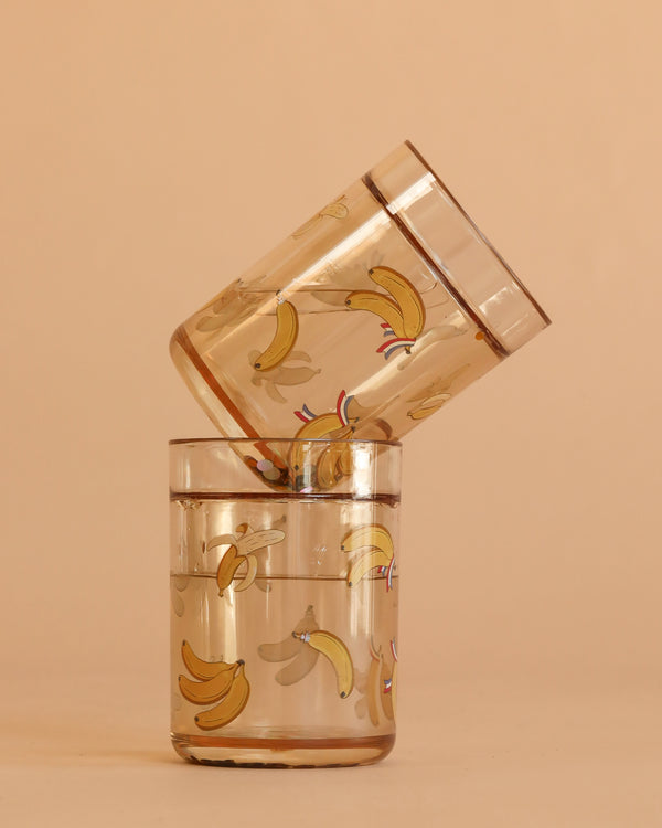 The Konges Sloejd Glitter Cups - Let's Go Bananas (2 Pack) feature playful banana illustrations, with one upright and filled, the other empty and horizontal. Their clear design and soft beige background make them ideal for fun kids' dinnerware collections.