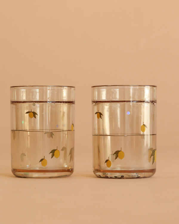 Two Konges Sloejd Glitter Cups - Lemon (2 Pack) filled with water, featuring yellow lemon and green leaf designs, sit side by side against a light beige background.