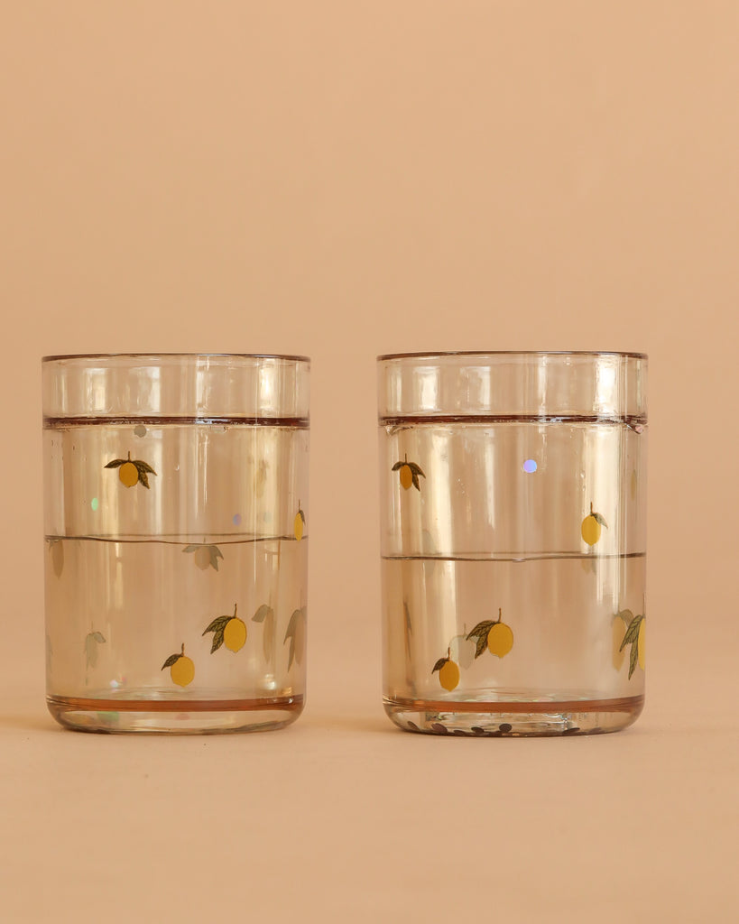 Two Konges Sloejd Glitter Cups - Lemon (2 Pack) filled with water, featuring yellow lemon and green leaf designs, sit side by side against a light beige background.
