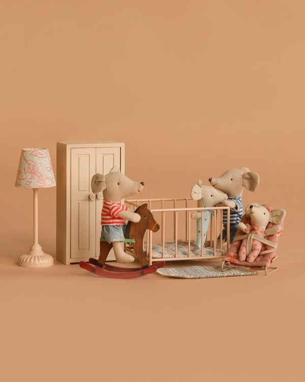 The Maileg Nursery Set with Brothers and Sisters features three mice—one on a rocking horse, one in a crib, and one in a chair—plus baby mouse accessories like a rug, lamp, and wardrobe arranged on a beige background.