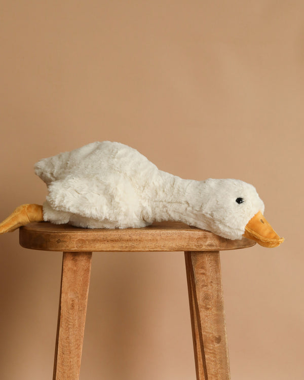 The Steiff XL Gilda Goose, with a yellow beak and feet, lies stretched out on a wooden stool against a beige background—an irresistibly huggable plush for any collection.