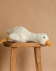 The Steiff XL Gilda Goose, with a yellow beak and feet, lies stretched out on a wooden stool against a beige background—an irresistibly huggable plush for any collection.