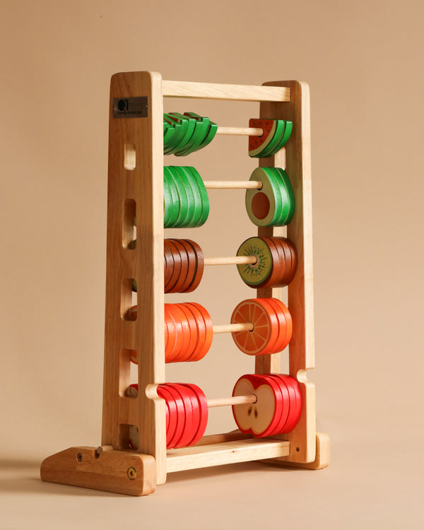 A Fruitful Abacus stands against a beige background, showcasing vibrant fruit-shaped beads on five horizontal rods. The beads represent orange slices, red apples, green leaves, kiwis, and oranges. This educational tool brings a playful twist to honing counting skills.