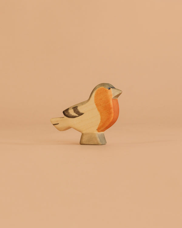 The Handmade Holzwald Finch Bird, featuring an orange breast, beige body, and dark accents, stands on a flat surface against a peach background—ideal for anyone seeking sustainable toys.