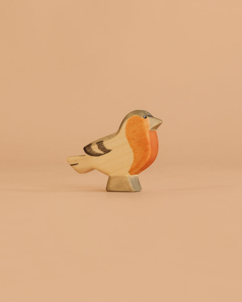 The Handmade Holzwald Finch Bird, featuring an orange breast, beige body, and dark accents, stands on a flat surface against a peach background—ideal for anyone seeking sustainable toys.