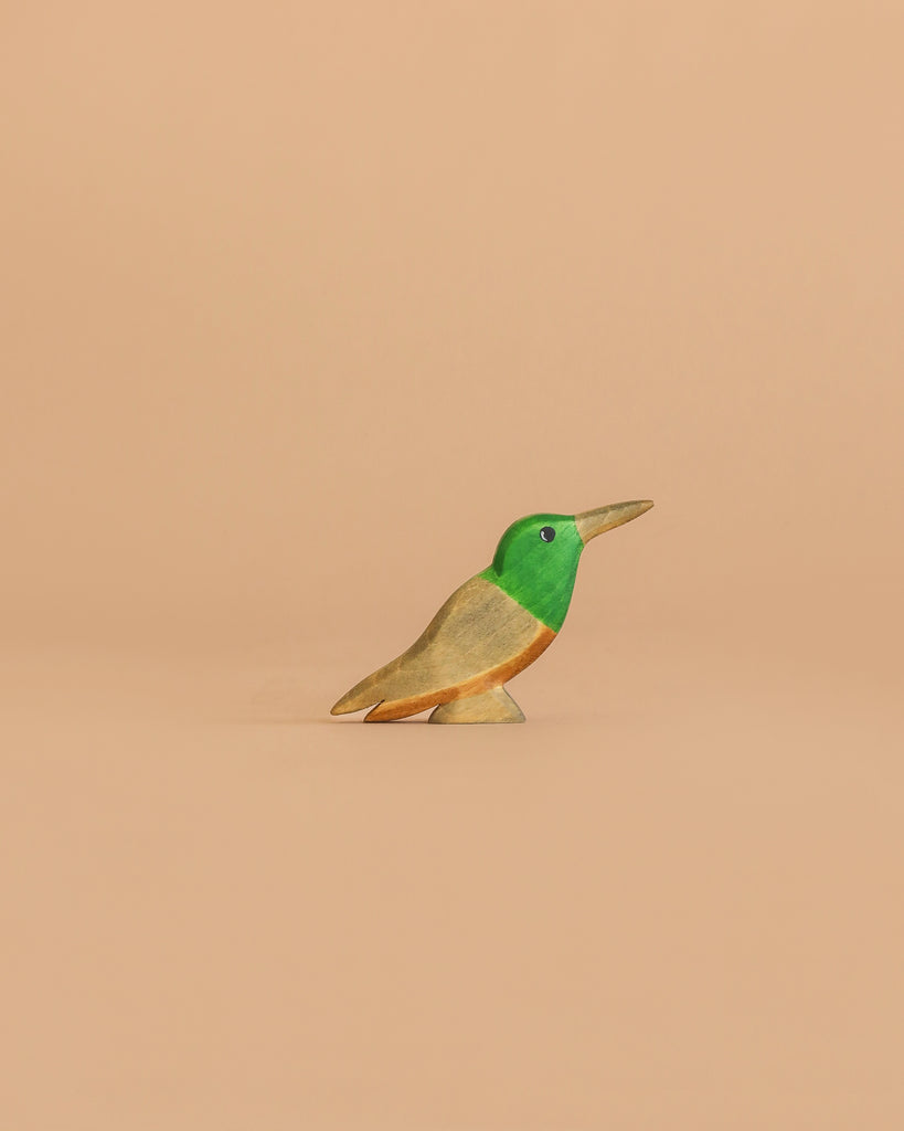 The Handmade Holzwald Hummingbird, a small wooden figurine painted green, yellow, and brown, is centered against a plain beige background, highlighting the charm of sustainable toys.