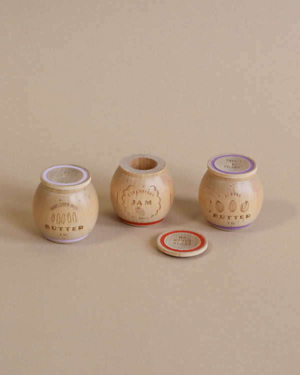 Three wooden containers with lids labeled "Milton & Goose Jam & Nut Butter Pretend Play Food" displayed against a beige background, with the lids resting beside them.