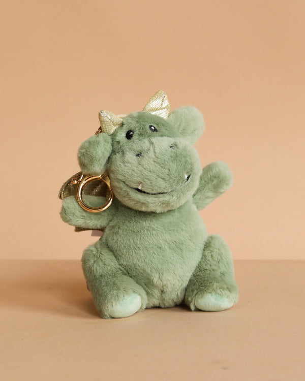 The Mon Ami Dax Dragon Keychain is a small green plush dragon with gold fabric horns and a smiling face, sitting upright against a beige background—perfect as a cute bag charm or accessory.