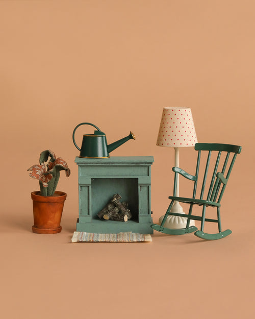 The Maileg Cozy Green Room Set features a miniature green fireplace with logs, watering can, potted plant, striped rug, vintage floor lamp, and rocking chair—all arranged on a beige background.