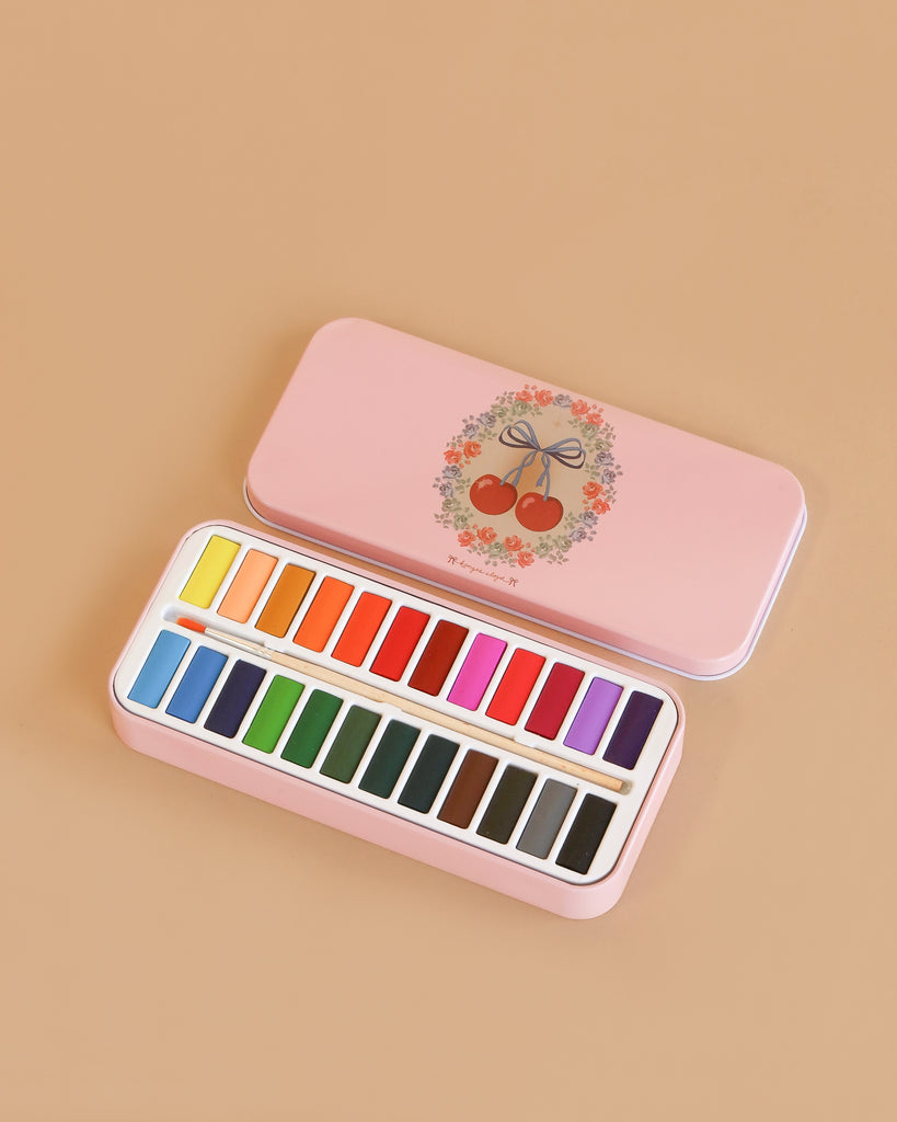 The Konges Sloejd Water Colors Paint Set - Cherry Bow features a pink tin box with cherry and floral lid design, holding neatly arranged colorful rectangular watercolors in two rows, displayed on a beige surface.