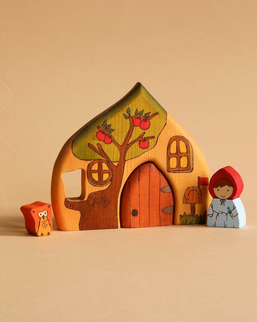 The Handmade Wooden Cottage set features a charming, hand-painted wooden cottage with tree detailing, a small figure resembling Little Red Riding Hood, and an owl figure. The cottage boasts arched windows and a central door, decorated with brown and green accents. This delightful toy set is ideal for play or as decorative decor in any room.