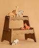 A wooden step stool displays the Ostheimer Handmade Wooden Safari Animals Set—colorful giraffe, zebra, lion, tiger with cub on top, and two elephants below—all handcrafted toys set against a beige background.