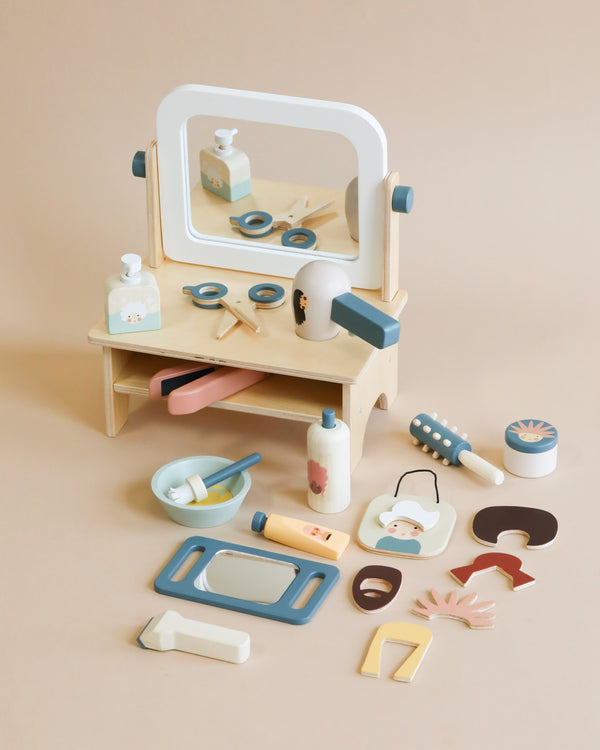 A wooden toy Hair Salon set with a mirror and various toy accessories including a hairdryer, brushes, and hair styling products, all laid out on a beige background.