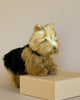 A realistic, Yorkshire Terrier Dog Stuffed Animal from HANSA plush animals with tan and black fur stands on a beige rectangular block against a neutral background. This hand-sewn animal has small, round eyes and a cute expression, mimicking a small breed like a Yorkshire Terrier.