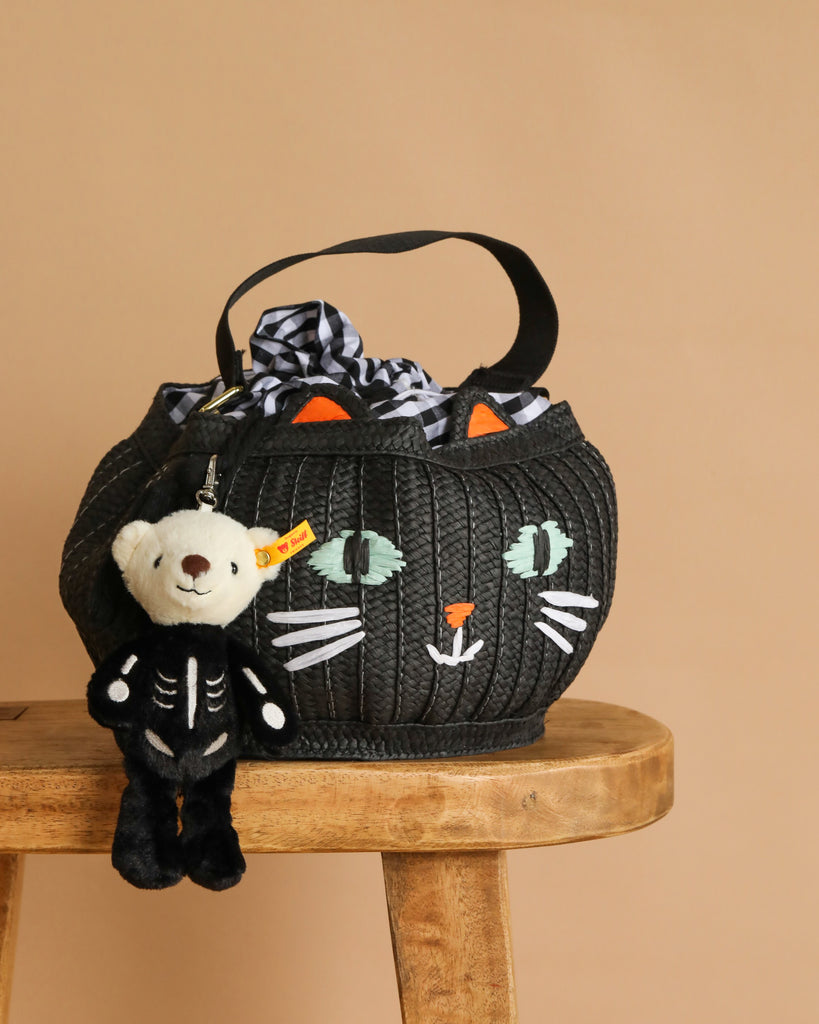 A Trick-or-Treat Basket with Steiff Mr. Bones Teddy Keychain—featuring a black cat’s face with orange ears and painted whiskers, and a skeleton-costumed bear keychain—sits on a wooden stool, perfect for Halloween treats!.