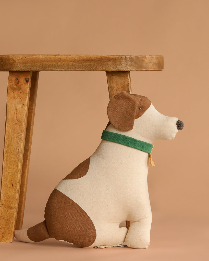 The Dog Cushion - Cooper, a plush dog-shaped pillow with brown and white patches and a green collar, sits upright next to a wooden stool against a beige background.