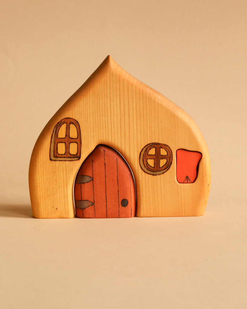 A small, charming Handmade Wooden Cottage stands against a beige background. Crafted in Ukraine, this hand-painted wooden structure features a pointed roof, a round arched door, circular and arched windows with simple details, and an orange triangular accent on one side.