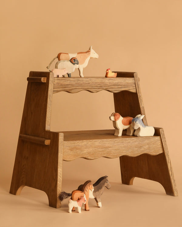 An Ostheimer Handmade Wooden Farm Animals Set, featuring horses, a sheep, and a chicken, is displayed on and around a wooden two-step stool against a beige background.