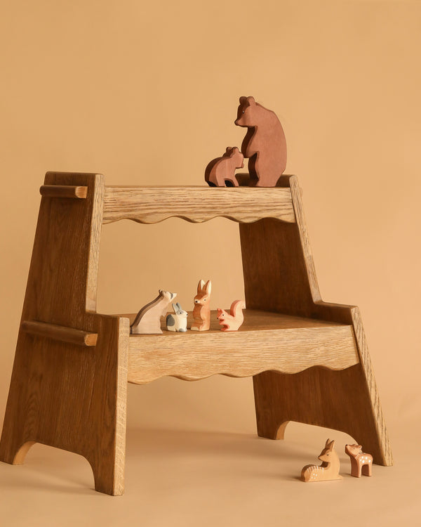 An Ostheimer Handmade Wooden Woodland Animals Set, featuring bears, a rabbit, a squirrel, a fox, and a deer, is displayed on a wooden step stool against a beige background.