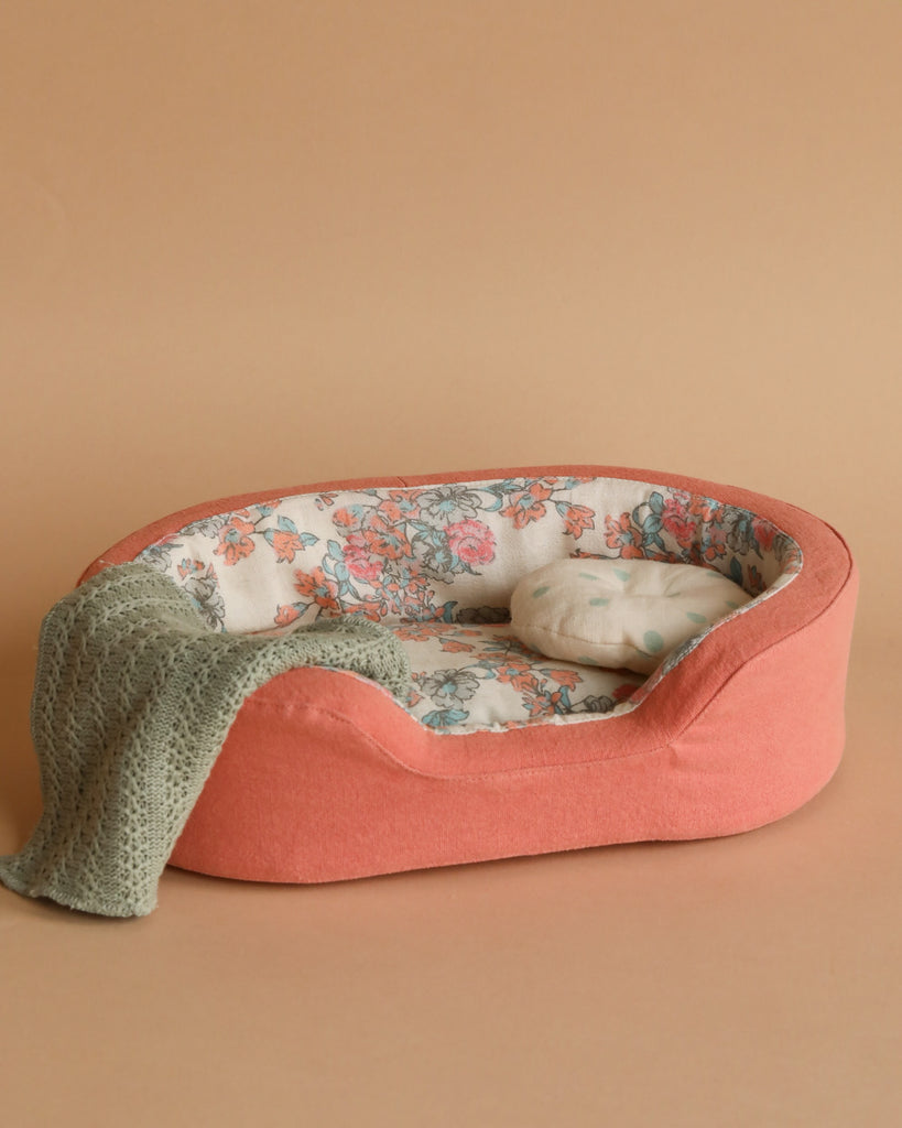 The Maileg Small Cozy Bed - Coral is an inviting, oval-shaped pet bed with a plush, coral-colored exterior and a charming floral-patterned interior. It comes with an adorable matching pillow and features a soft blanket tastefully draped over the side, all set against a beige backdrop.