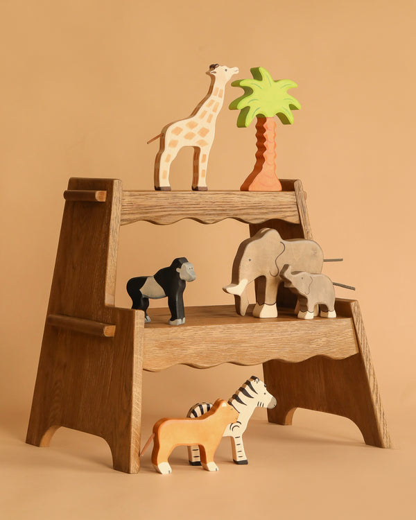 The Holztiger Handmade Wooden Safari Animals Set features a giraffe, gorilla, elephant, zebra, tiger, and a palm tree on a wooden step stool against a beige backdrop—perfect for imaginative play.