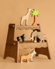 The Holztiger Handmade Wooden Safari Animals Set features a giraffe, gorilla, elephant, zebra, tiger, and a palm tree on a wooden step stool against a beige backdrop—perfect for imaginative play.
