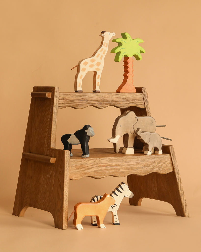 The Holztiger Handmade Wooden Safari Animals Set features a giraffe, gorilla, elephant, zebra, tiger, and a palm tree on a wooden step stool against a beige backdrop—perfect for imaginative play.